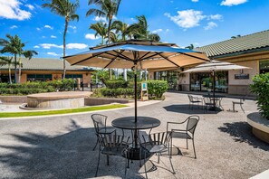 An outdoor patio area with round tables and chairs, surrounded by palm trees and a building with a tiled roof. - Spacious Townhome - Stunning Golf Course Views - Pool & Fitness- Vrkcv (Kamuela)