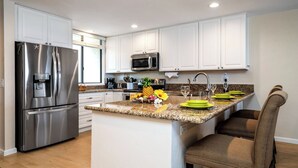 A modern kitchen with white cabinets, stainless steel appliances, a granite countertop, and a small island with seating. - No Stairs - AC - Fantastic Ocean & Golf Course Views! -vrkcv (Kailua-Kona)