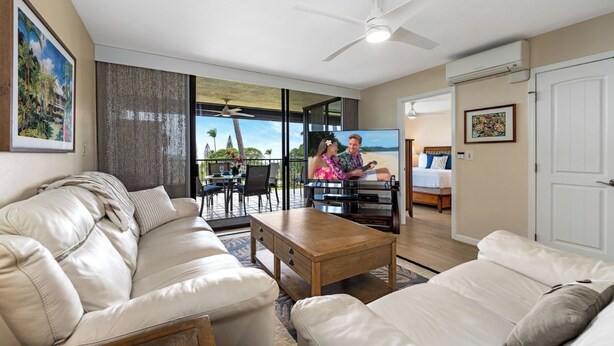A spacious living room with a sofa, coffee table, and a TV. There is a balcony with a dining area and a view of the outdoors. - No Stairs - AC - Fantastic Ocean & Golf Course Views! -vrkcv (Kailua-Kona)