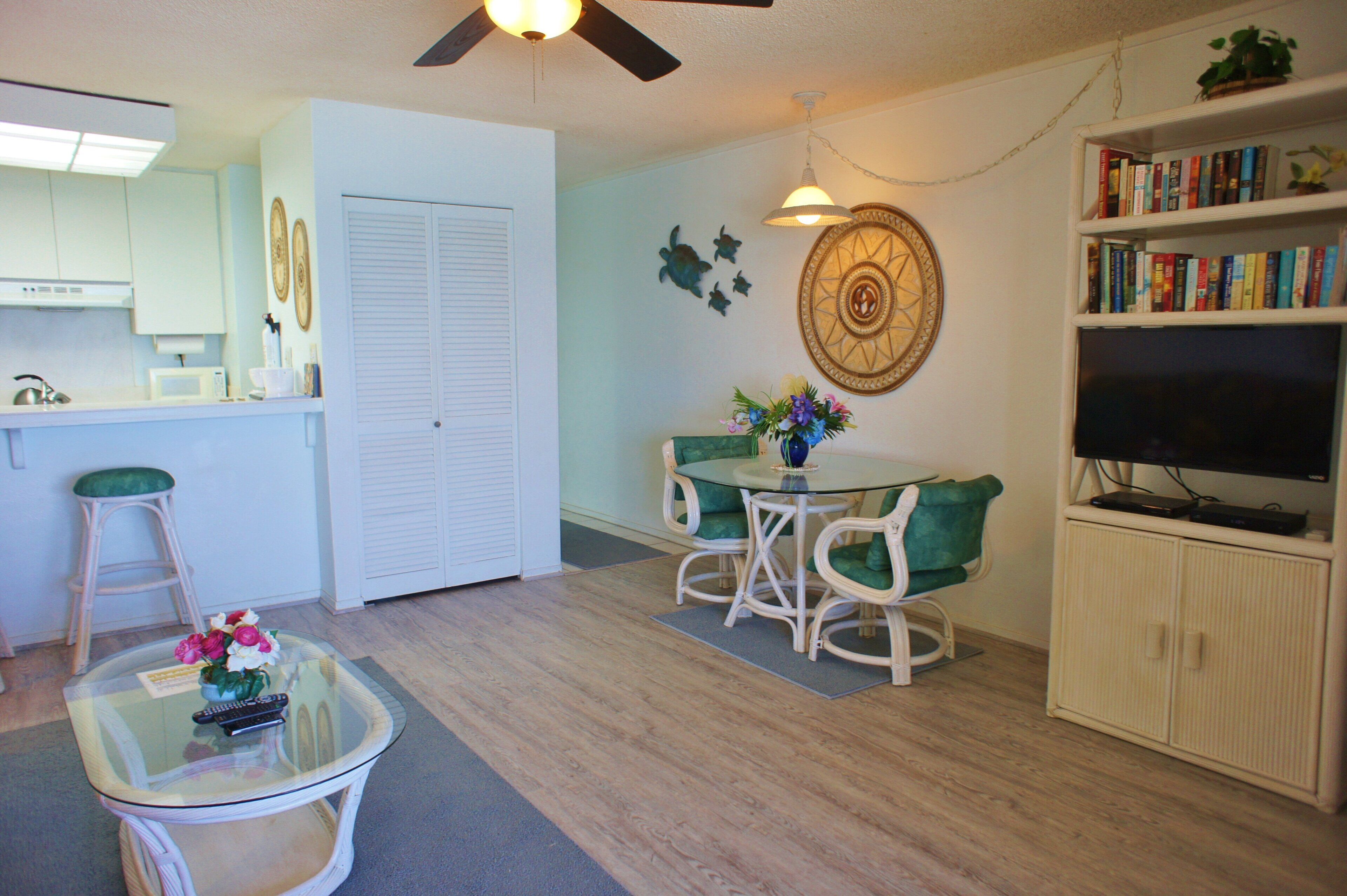 A living room with a glass coffee table, a dining table with chairs, a bookshelf, a television, and a ceiling fan.
