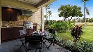 Une cuisine extérieure équipée d’un barbecue, d’une table à manger et de chaises, le tout entouré d’une végétation luxuriante et de palmiers.