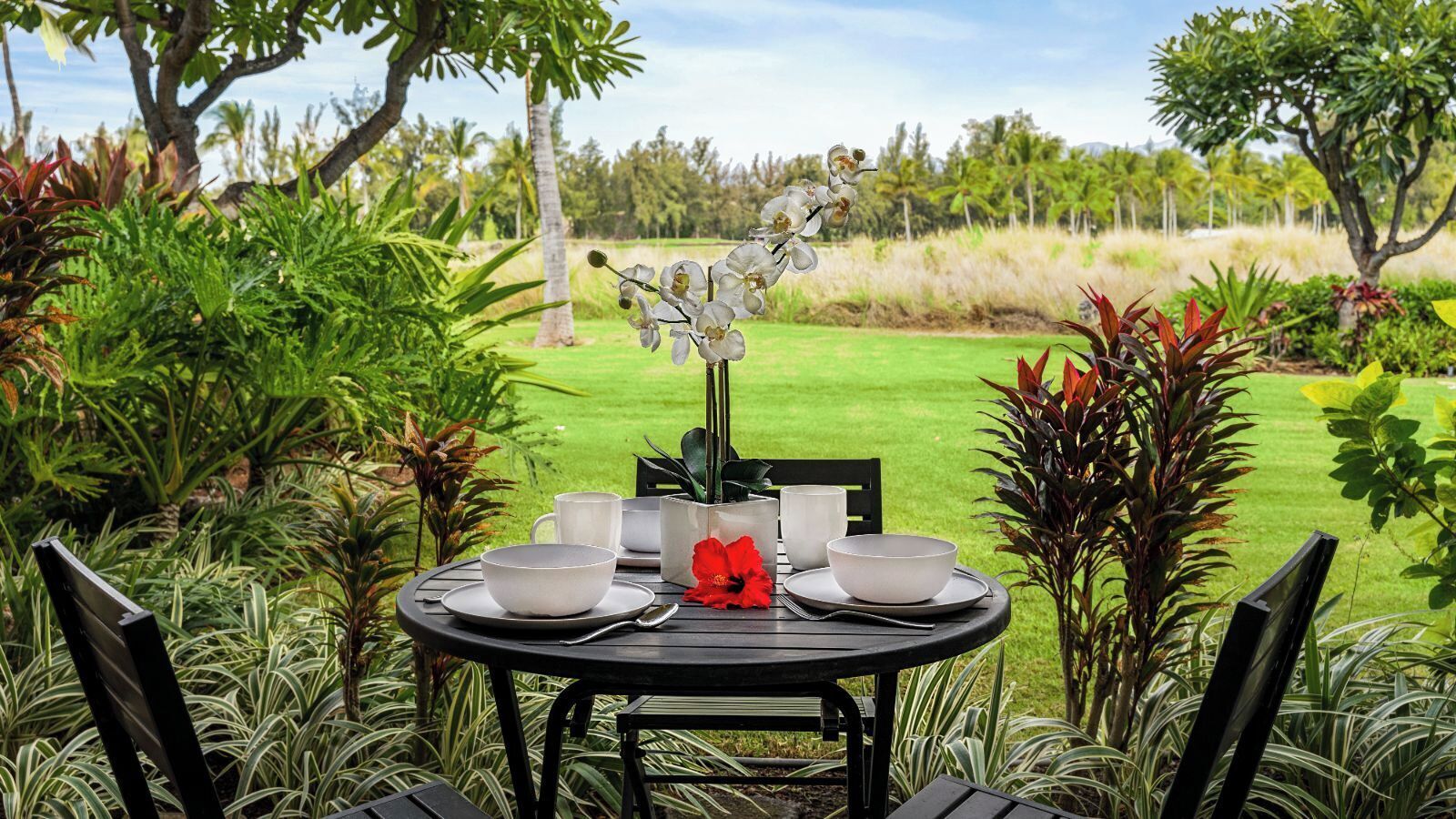 A round table set for two with white bowls, cups, and a red flower centerpiece, surrounded by lush greenery and palm trees.
