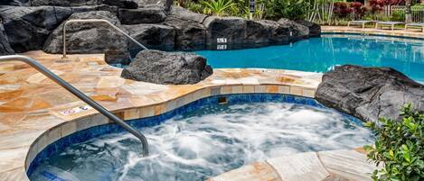 A hot tub with a stone surround, a pool with clear blue water, and a tropical garden setting.
