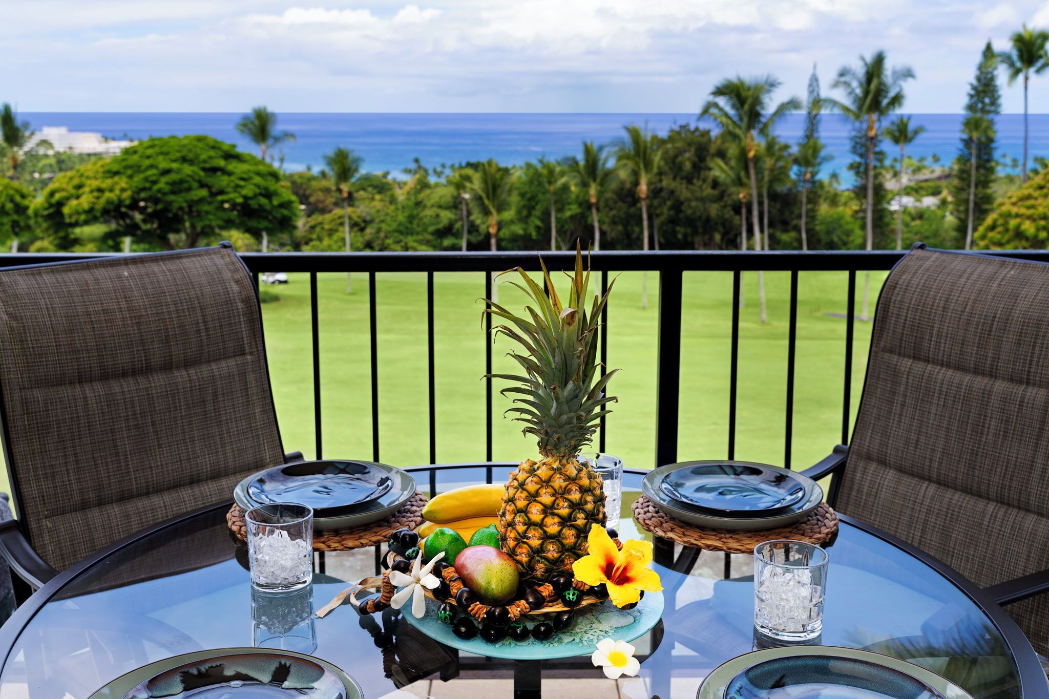 A table with a pineapple, bananas, and other fruits, set on a balcony with a view of the ocean and palm trees.