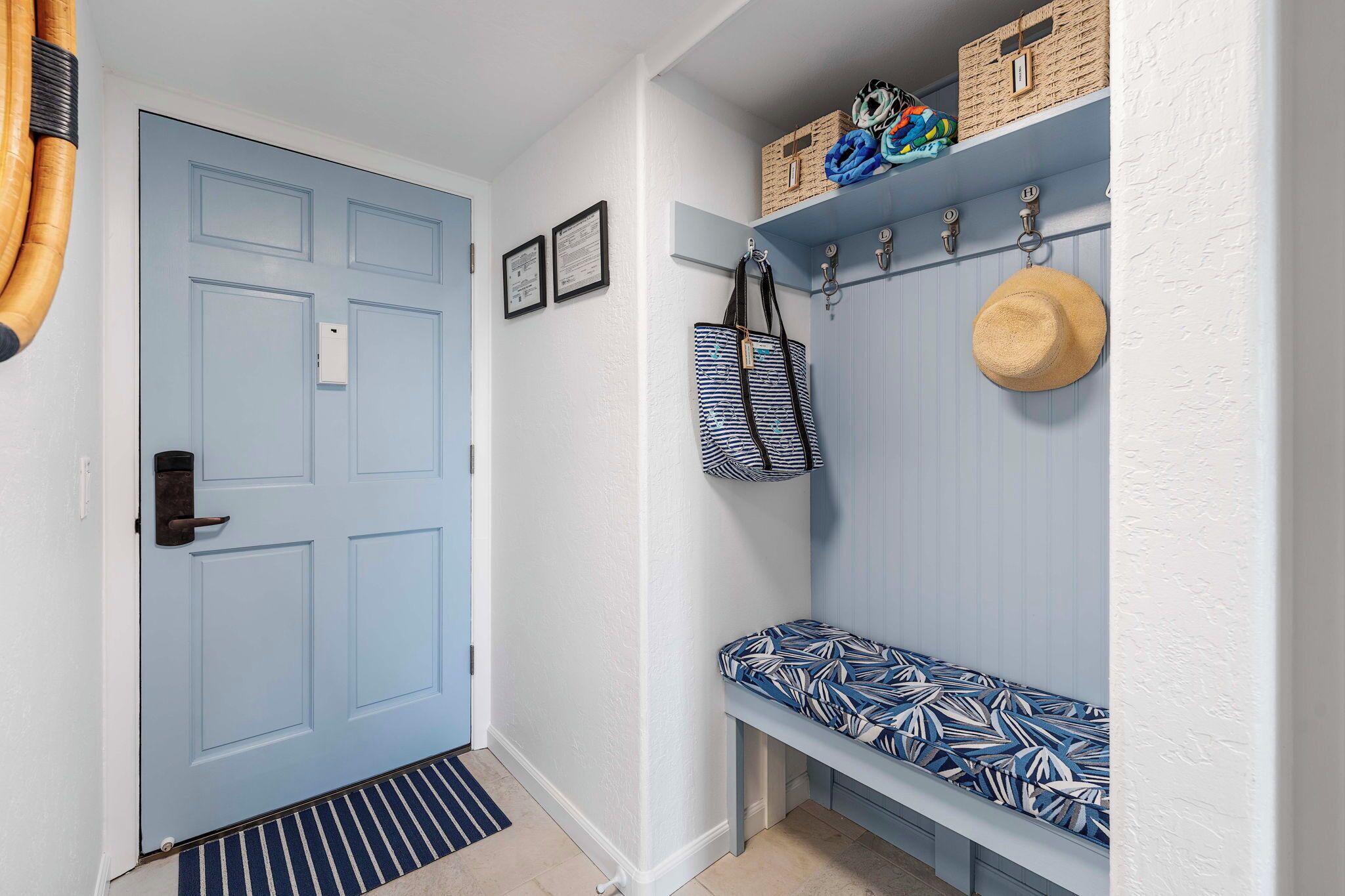 A hallway with a light blue door, a bench with patterned cushions, and a wall-mounted coat rack with hats and bags.