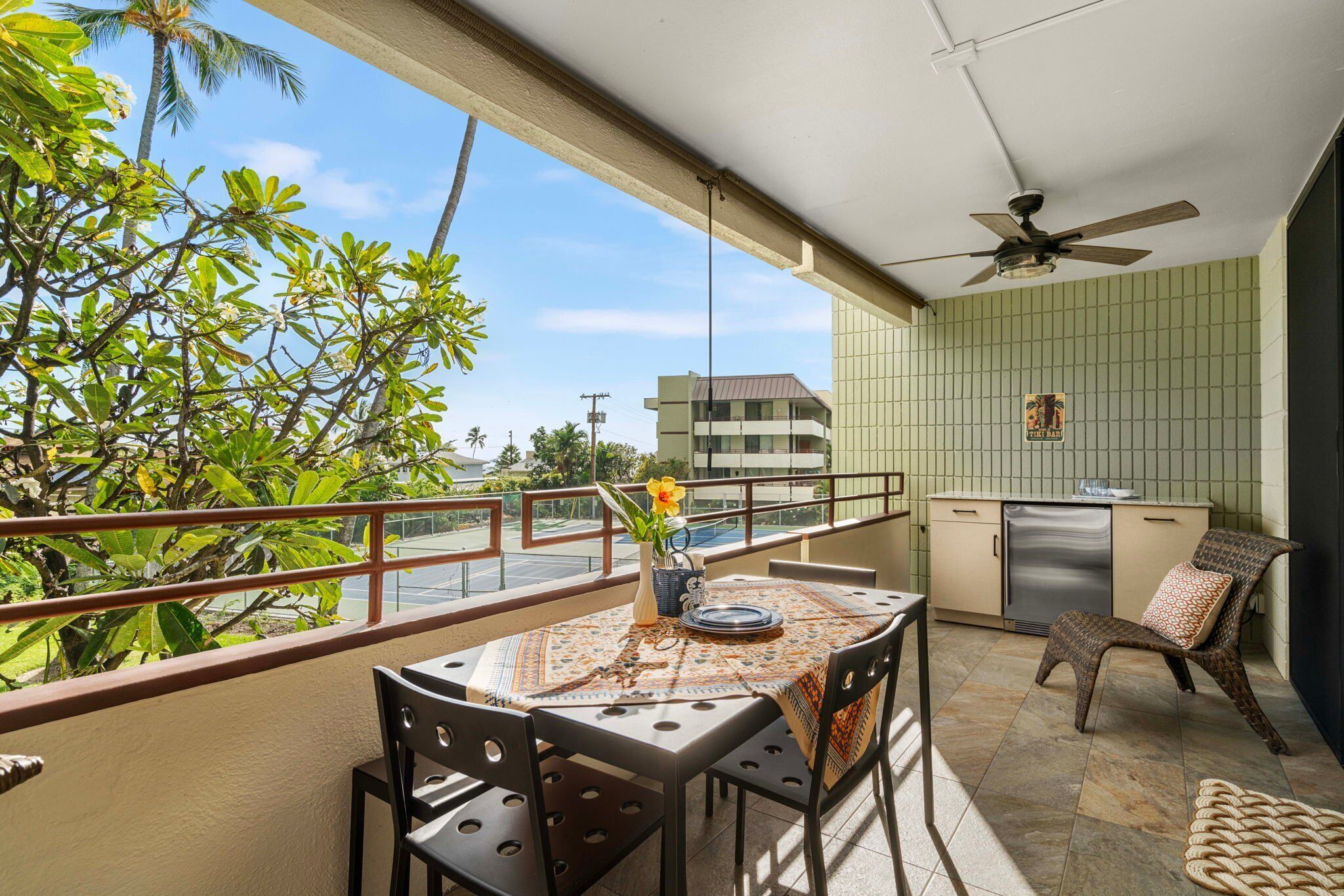 A balcony with a table, chairs, a ceiling fan, and a view of greenery and buildings.