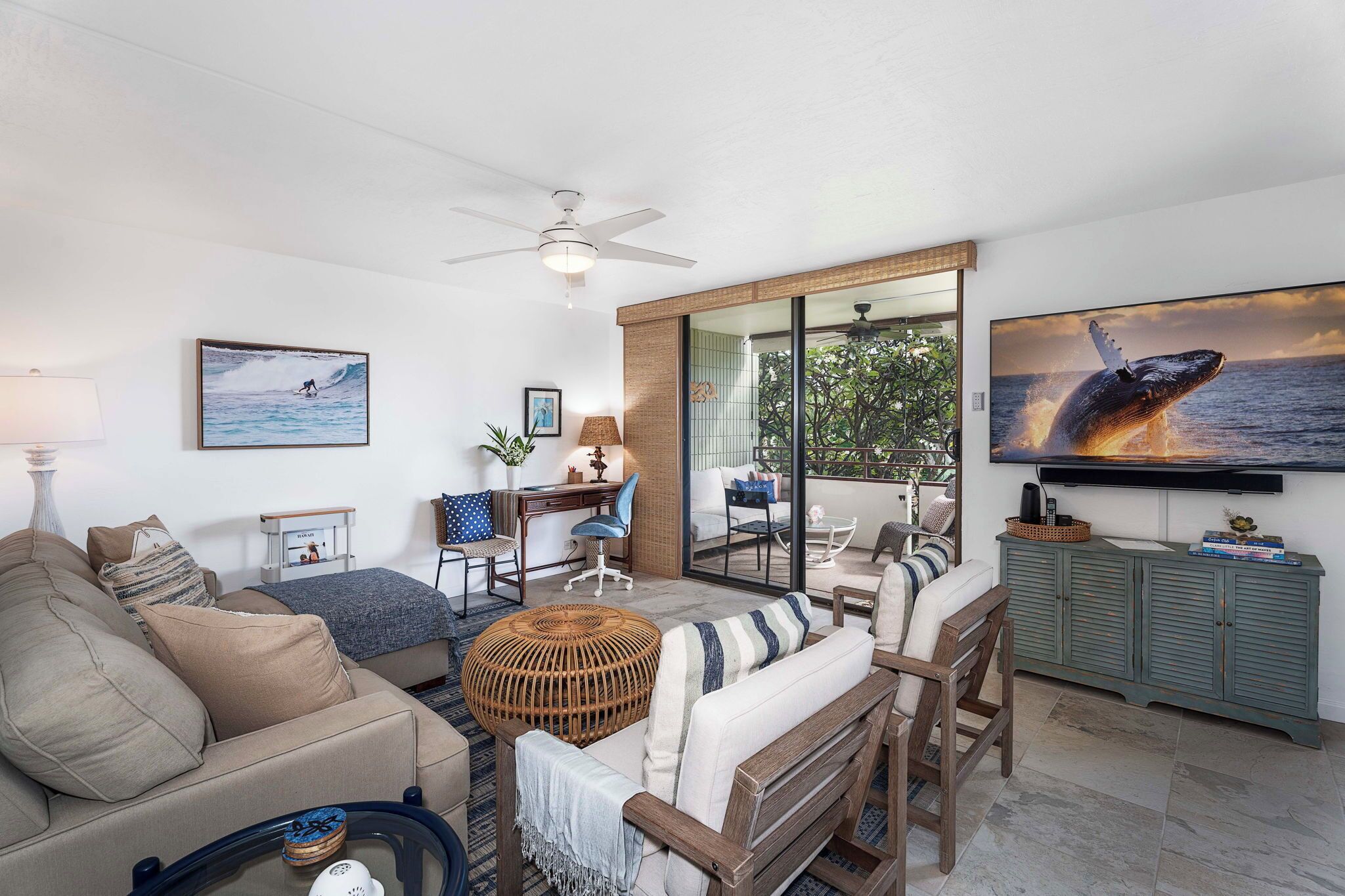 A spacious living area with a sofa, armchairs, a coffee table, and a television mounted on the wall. There is a sliding glass door leading to an outdoor area with a view of the ocean.