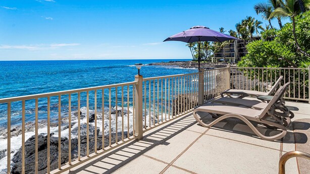 A beachside terrace with a view of the ocean, a lounge chair, and a purple umbrella.