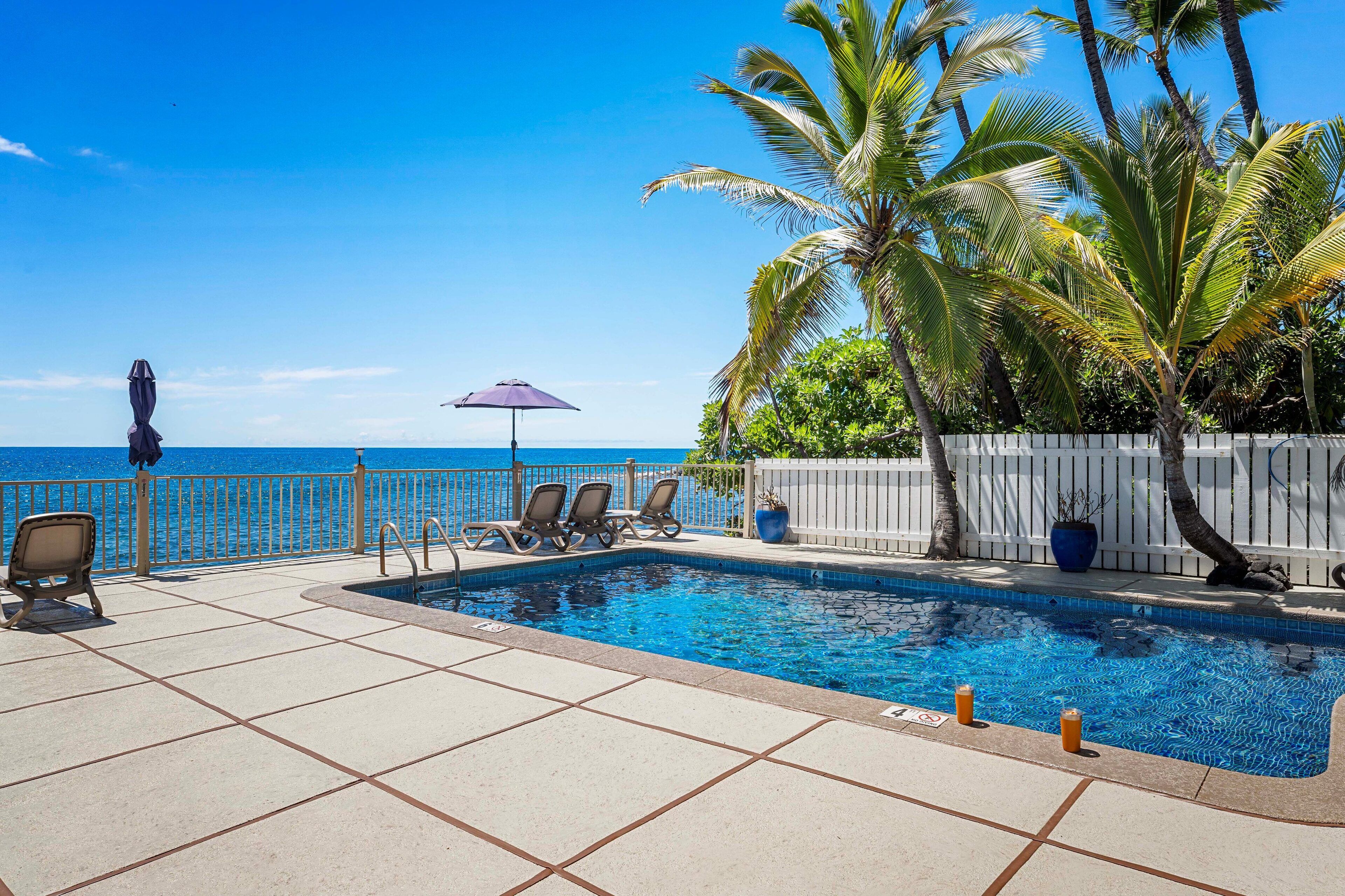 A pool area with a view of the ocean, lounge chairs, and a white fence.