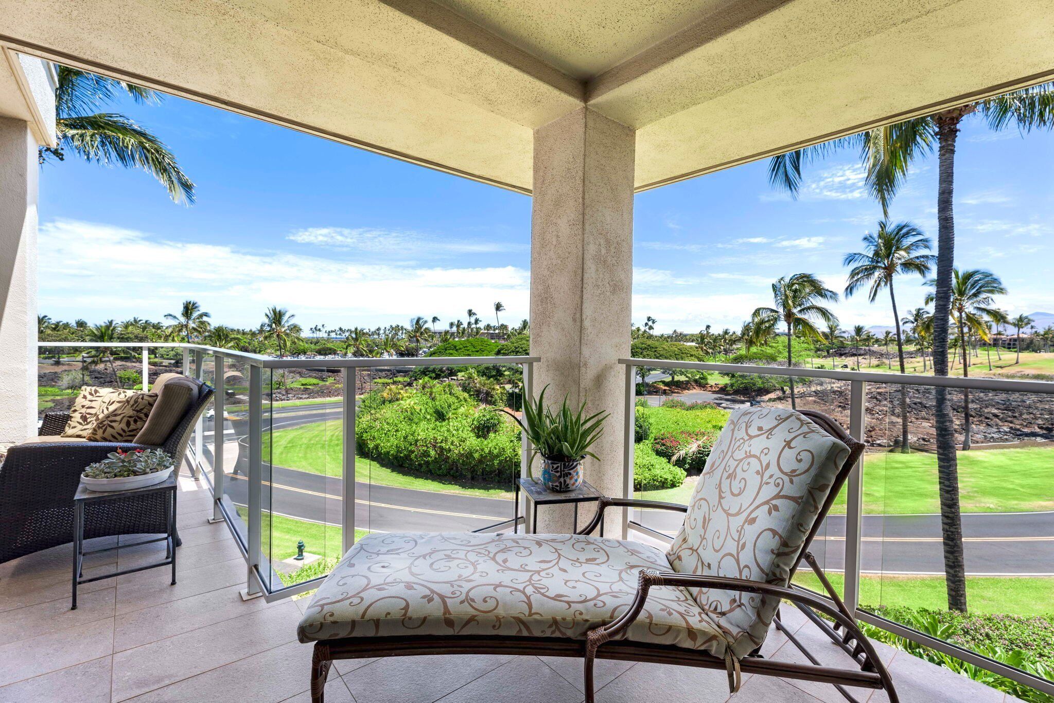 A covered balcony with two chairs and a table, overlooking a lush landscape with palm trees and a road.