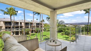A balcony with a sofa and a small table, overlooking a well-maintained garden and a building with multiple balconies. - Top Floor Rental - Ocean Views - Vrkcv (Waikoloa)