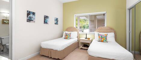 A bedroom with two beds, a window with blinds, a bedside table, and a small shelf.