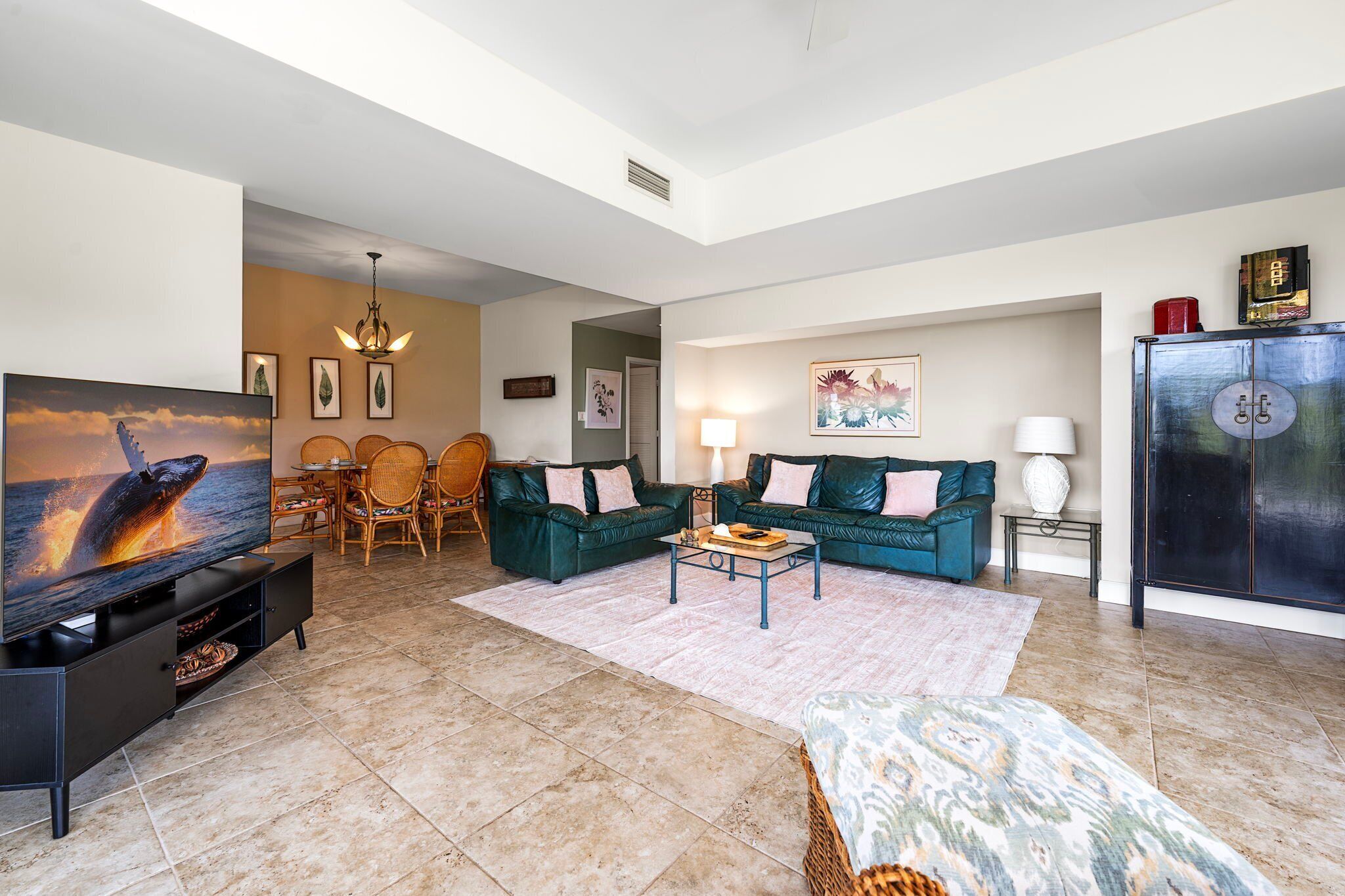 A spacious living room with a large flat-screen TV, a green sofa set, a coffee table, and a dining area with chairs and a table.