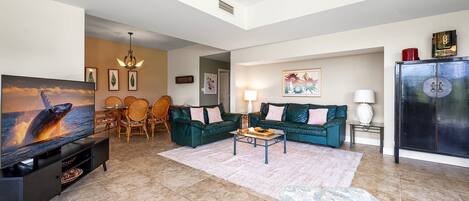 A spacious living room with a large flat-screen TV, a green sofa set, a coffee table, and a dining area with chairs and a table.