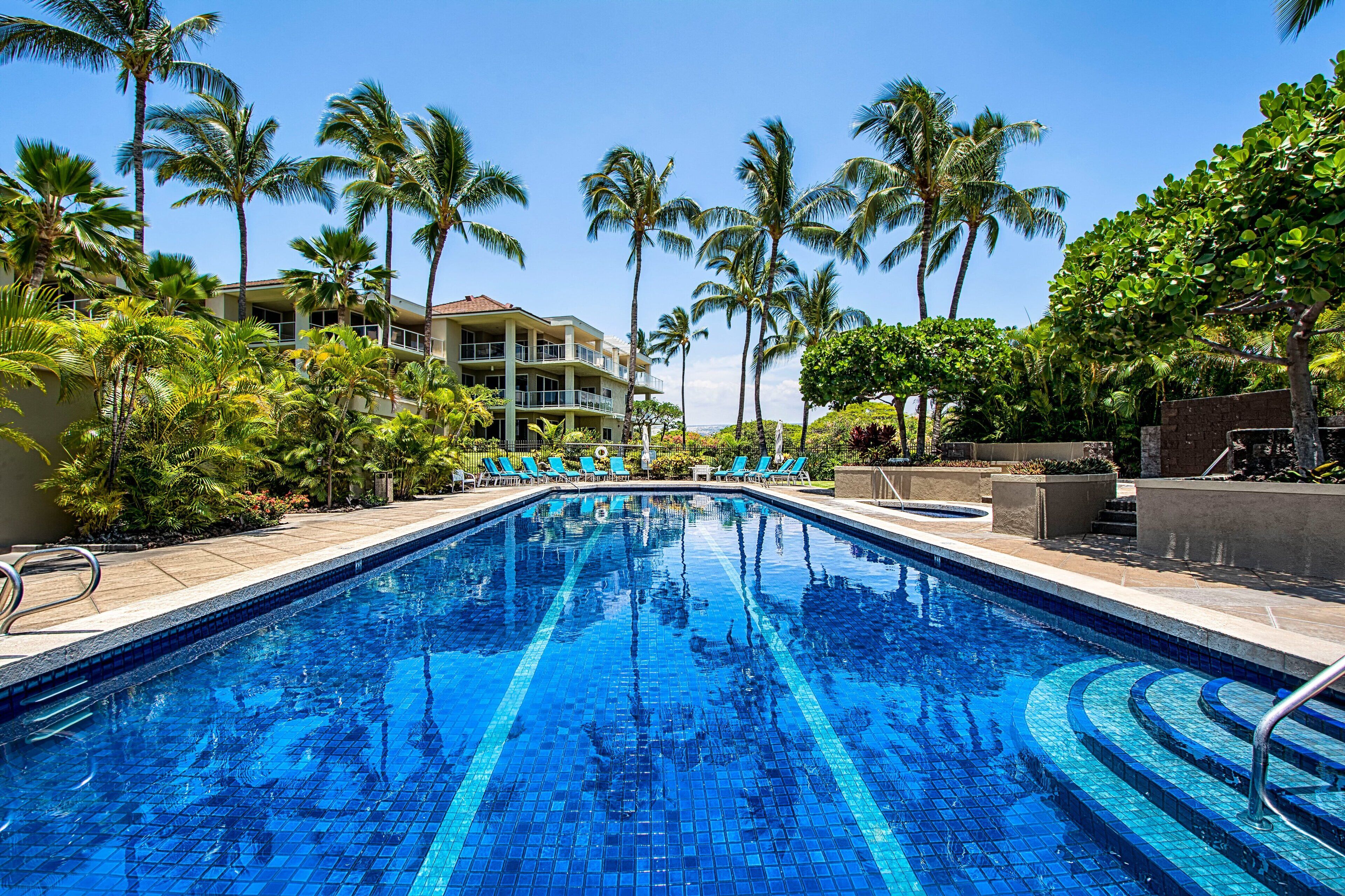 A swimming pool with clear blue water, surrounded by palm trees and a building with balconies.