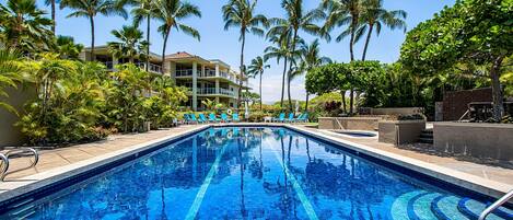 A swimming pool with clear blue water, surrounded by palm trees and a building with balconies.