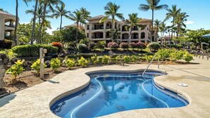 A swimming pool with a curved design, surrounded by palm trees and a landscaped garden. - Ground Floor - In-unit Central AC - Vrkcv (Waikoloa)