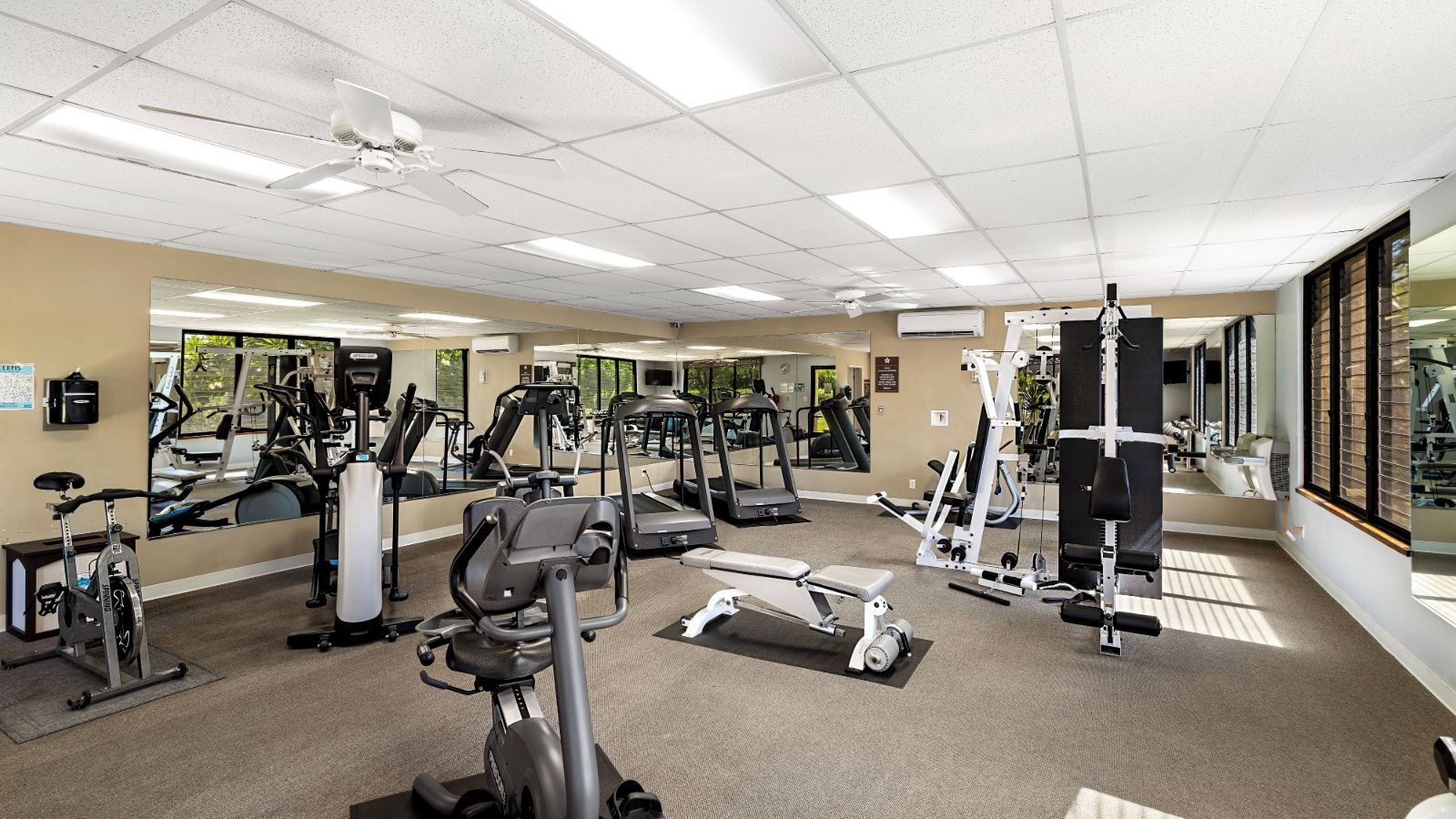 Un gimnasio con diferentes máquinas de ejercicio, amplias ventanas y un ventilador de techo.