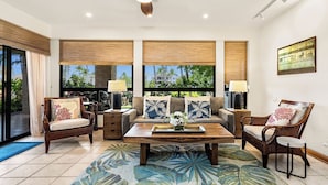 A spacious living room with a sofa, two armchairs, a coffee table, and a side table. Large windows with blinds provide views of palm trees and buildings. - Ground Floor - In-unit Central AC - Vrkcv (Waikoloa)