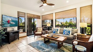 A living room with a television, sofa, coffee table, and a view of palm trees through large windows. - Ground Floor - In-unit Central AC - Vrkcv (Waikoloa)