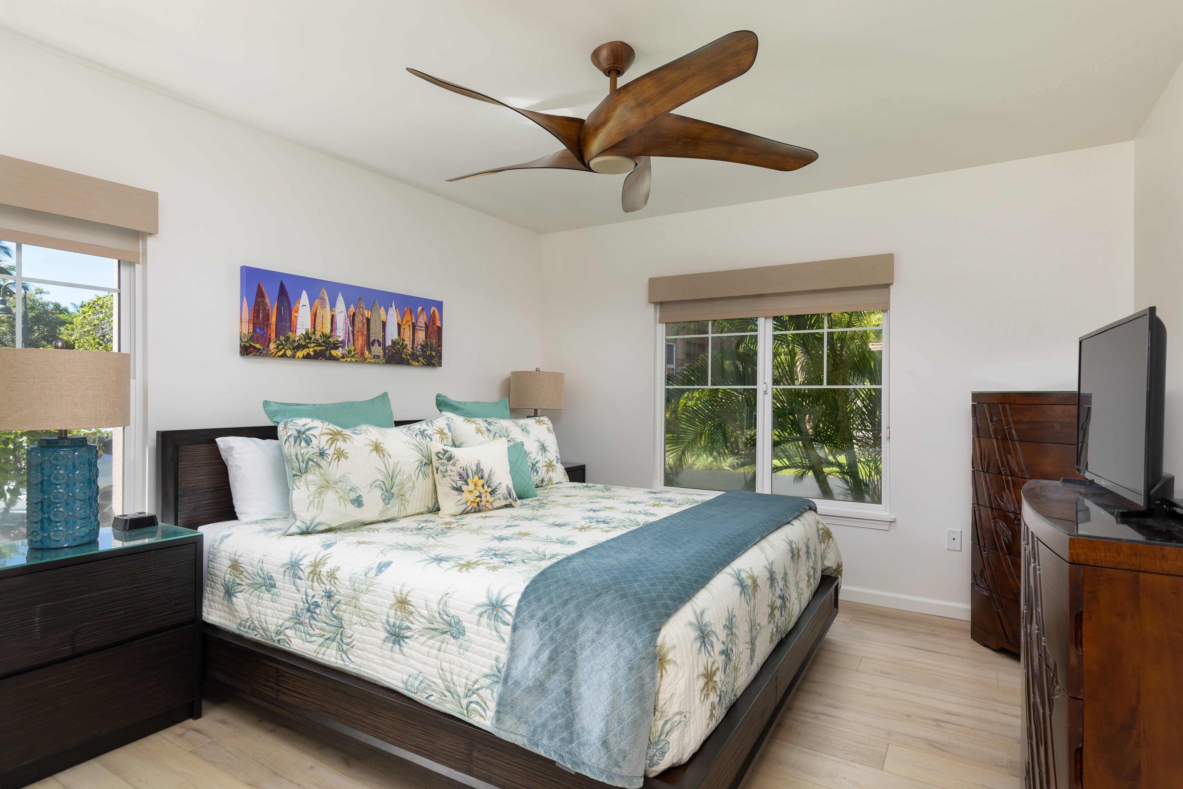 A bedroom with a bed, a ceiling fan, a window with a view of greenery, a painting on the wall, and a TV on a wooden cabinet.