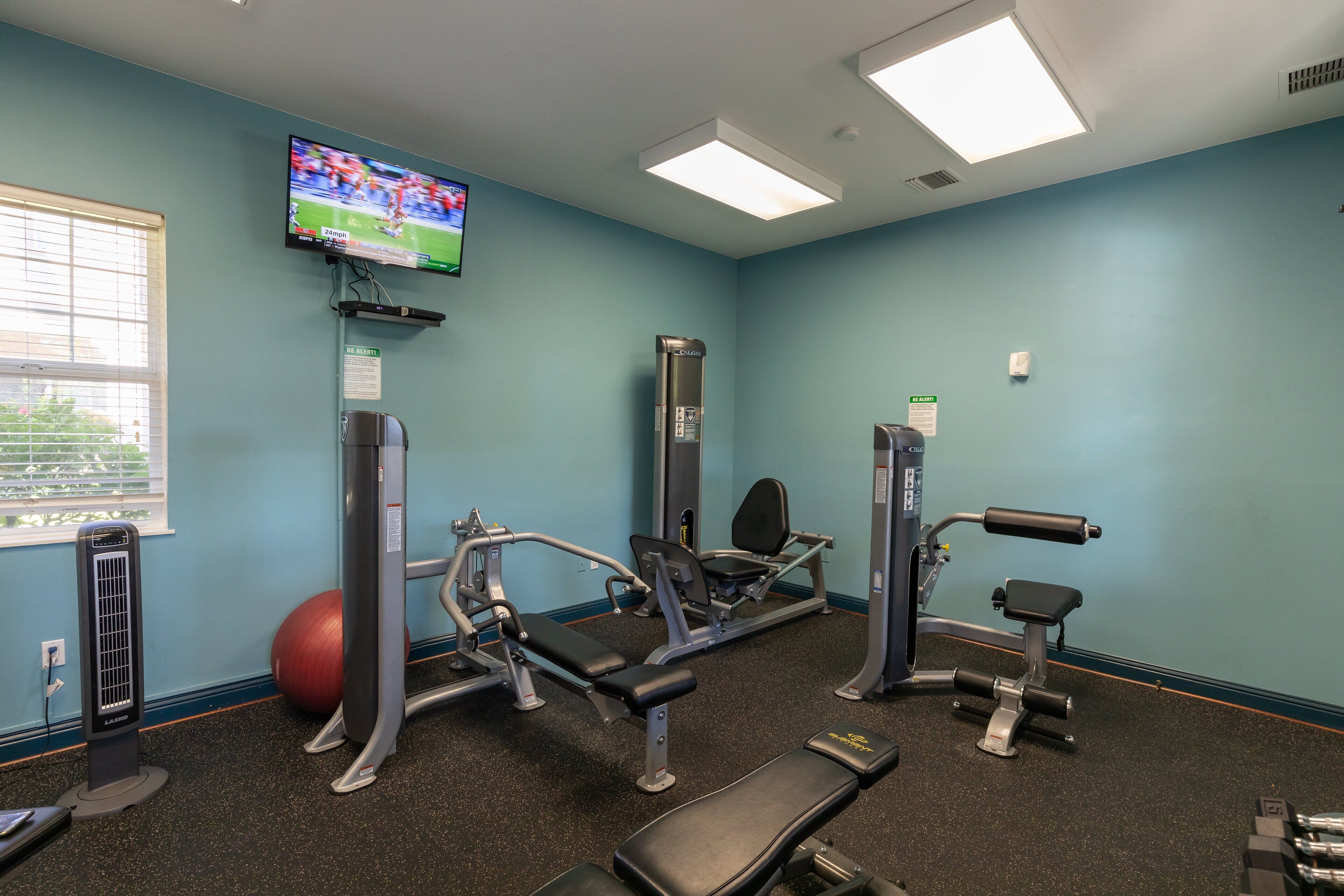A gym with exercise equipment, a television on the wall, and a window with blinds.