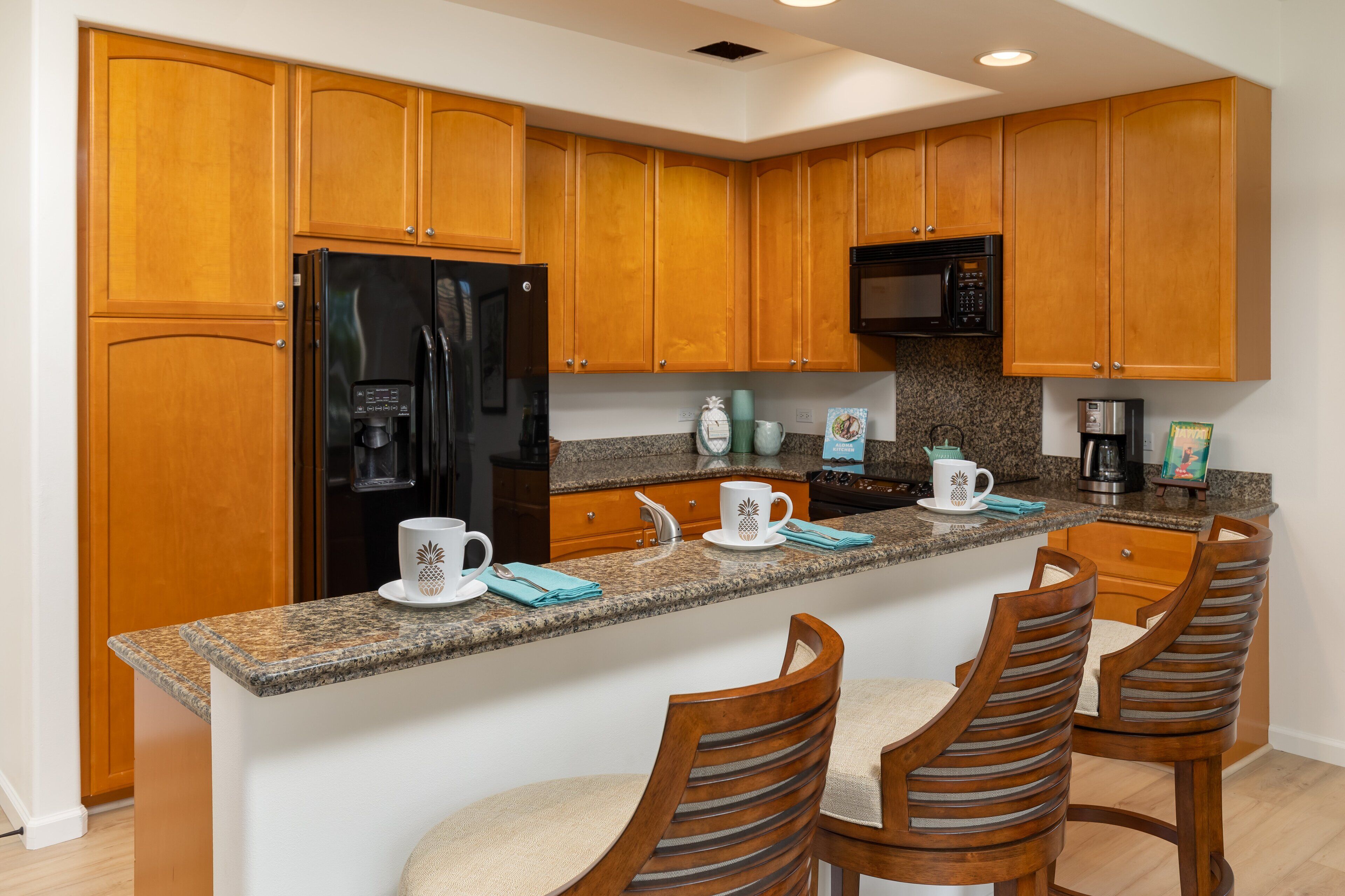 A kitchen with wooden cabinets, a granite countertop, a black refrigerator, a microwave, and a coffee maker. There are two wooden chairs with cushioned seats.