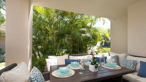 Un área de comedor al aire libre con una mesa de mimbre dispuesta para dos personas, rodeada de asientos acolchados y un exuberante follaje tropical.