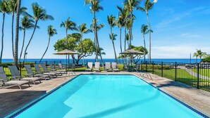 A swimming pool with lounge chairs and umbrellas, surrounded by palm trees and a view of the ocean.