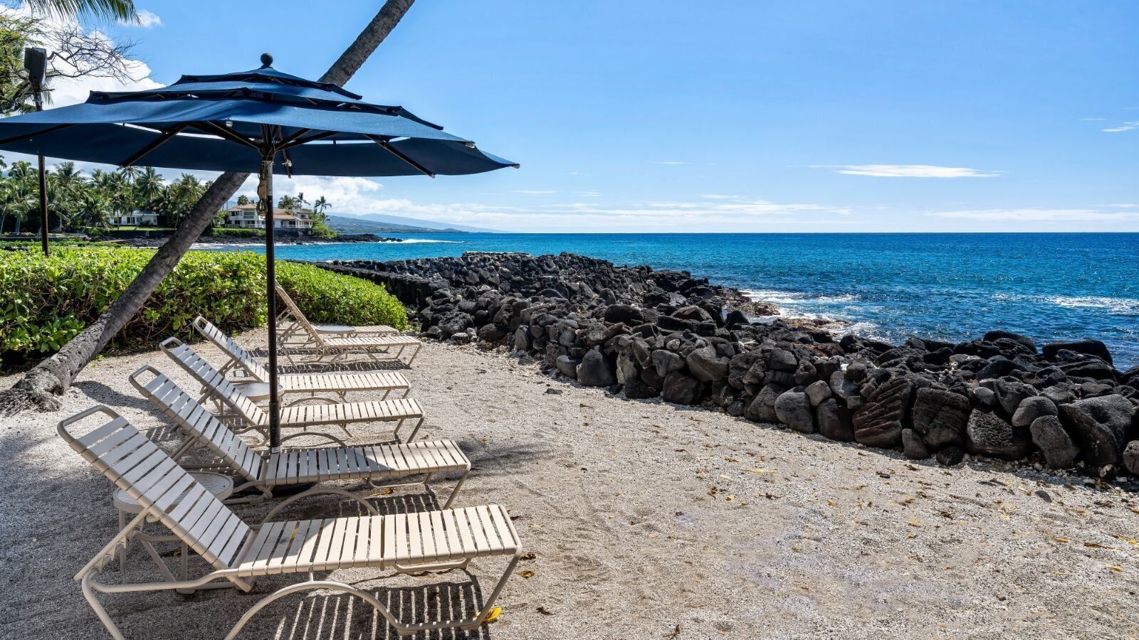 A beachside lounge with sun loungers and a large umbrella, overlooking a rocky shoreline and the ocean.