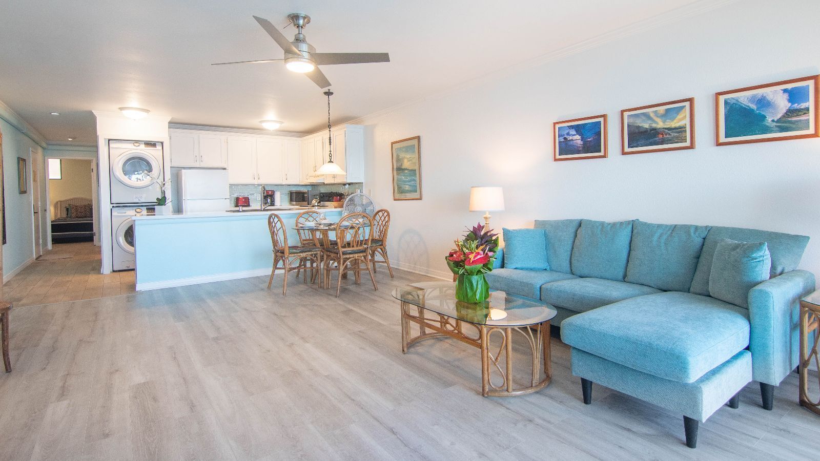 Una sala de estar con un sofá azul, una mesa de comedor con sillas, una zona de cocina con lavadora y un ventilador de techo.