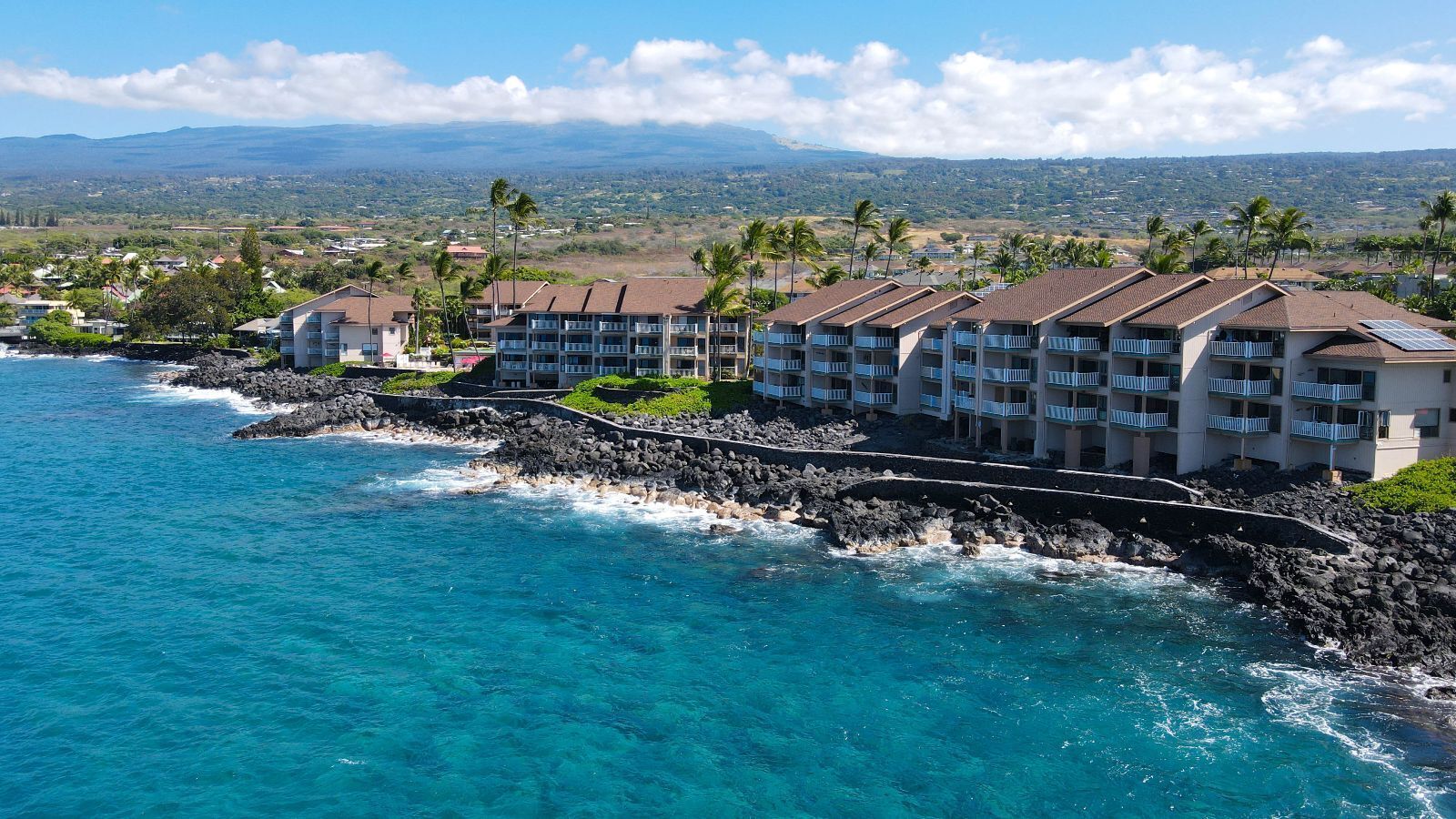 A coastal hotel with multiple balconies overlooking the ocean, a rocky shoreline, and a view of distant hills.