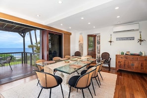 Un espace repas avec une table en verre, des chaises en osier, une terrasse en bois et une vue sur l’océan.