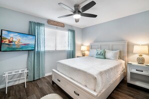 Une chambre à coucher avec un lit, un ventilateur de plafond, une télévision et une fenêtre avec des rideaux.
