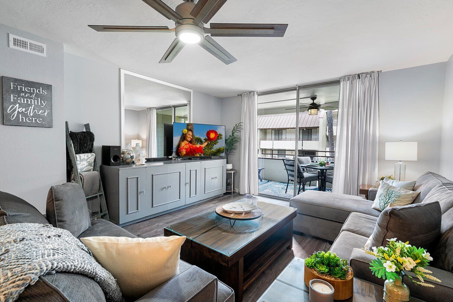 A modern living room with a grey sofa, a coffee table, and a TV on a cabinet. There's a balcony with a table and chairs, and a ceiling fan.