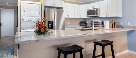 A modern kitchen with white cabinets, stainless steel appliances, a marble countertop, and two black bar stools.