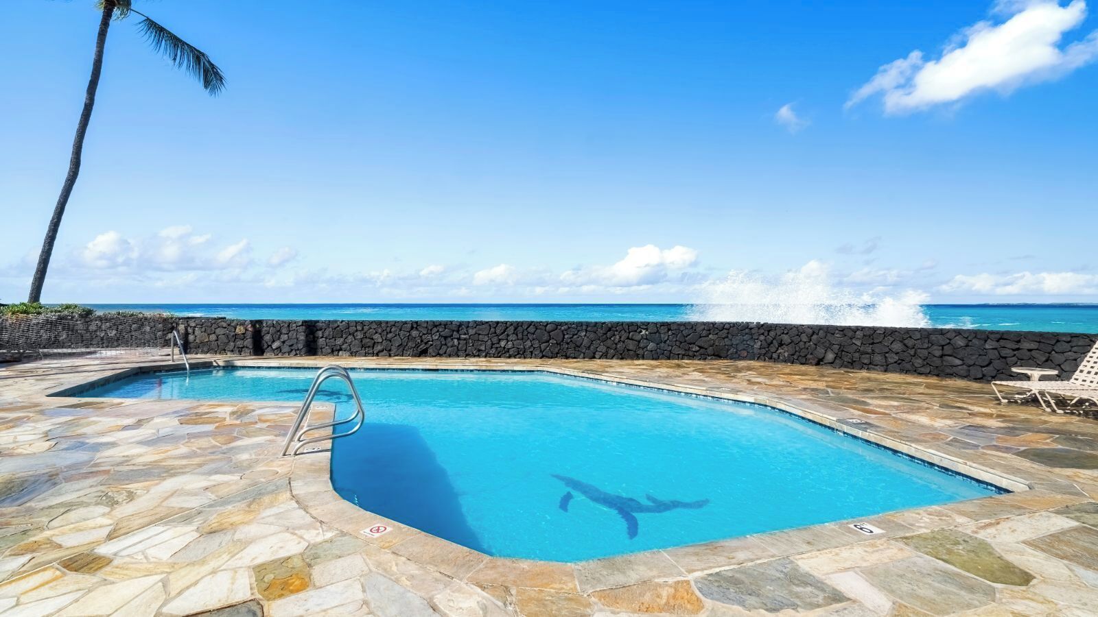En swimmingpool med stenterrasse, en livredderstol og udsigt til havet.