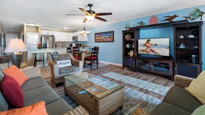 A living room with a television, wicker furniture, and a kitchen in the background. - Oceanfront - Free Concierge - Wifi - Top Floor - Vrkcv (Kailua-Kona)