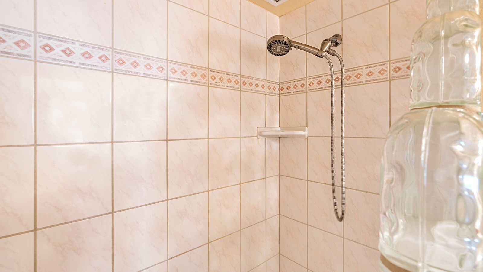 Une salle de bain avec des carreaux de couleur beige, une douche équipée d’un pommeau de douche amovible et une bouteille en verre transparent.