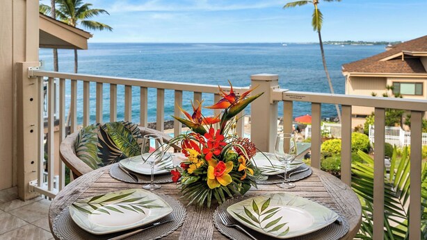 A table set for two with a tropical floral centerpiece, overlooking a coastal view. - Oceanfront - Free Concierge - Wifi - Top Floor - Vrkcv (Kailua-Kona)