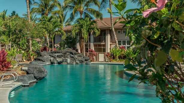 Une piscine d’un complexe hôtelier, avec une eau d’un bleu limpide, entourée de palmiers et bordée d’un bâtiment au toit recouvert de tuiles.