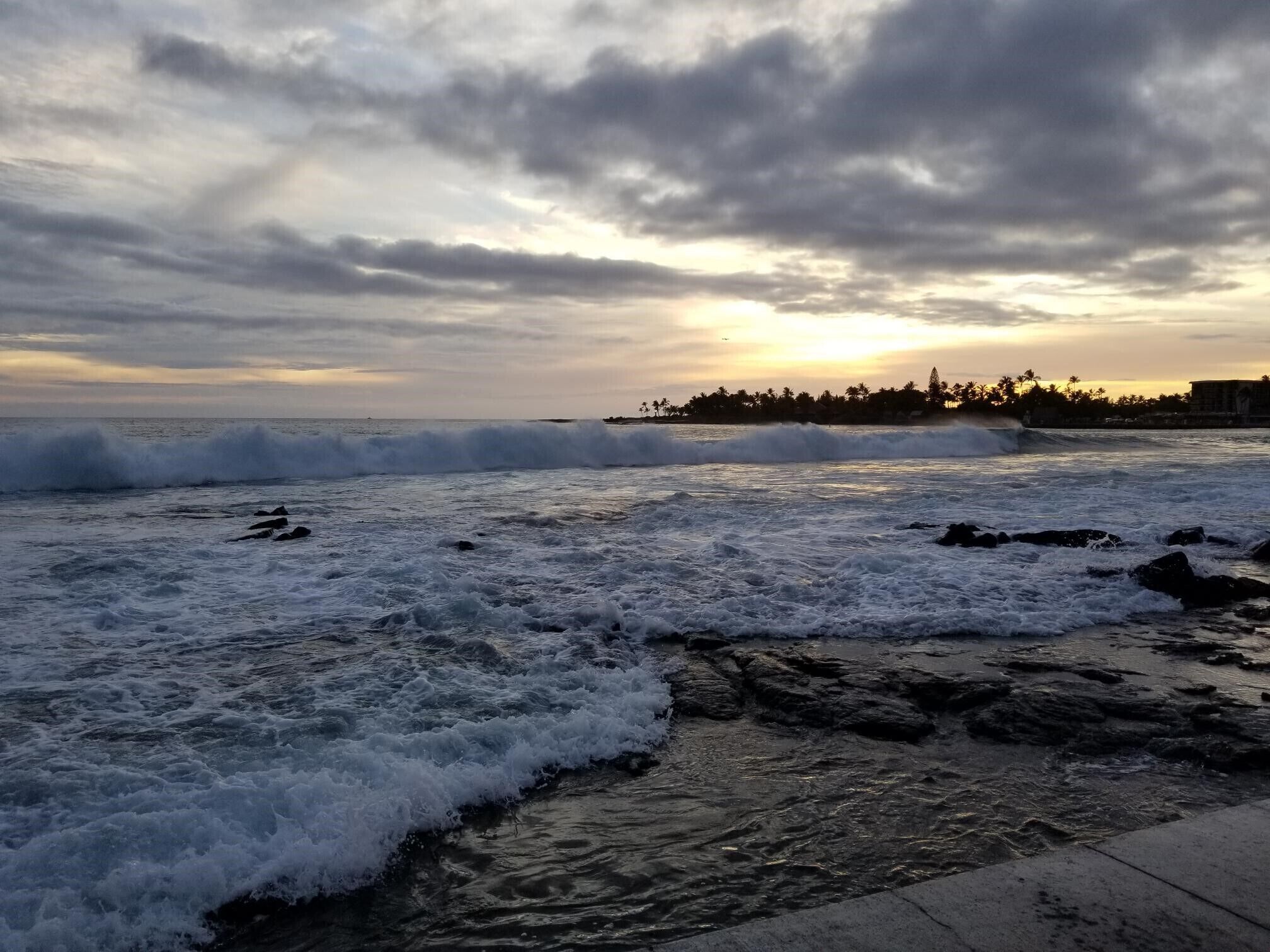 A coastal scene with waves crashing onto rocks and a distant shoreline with buildings and palm trees under a cloudy sky.
