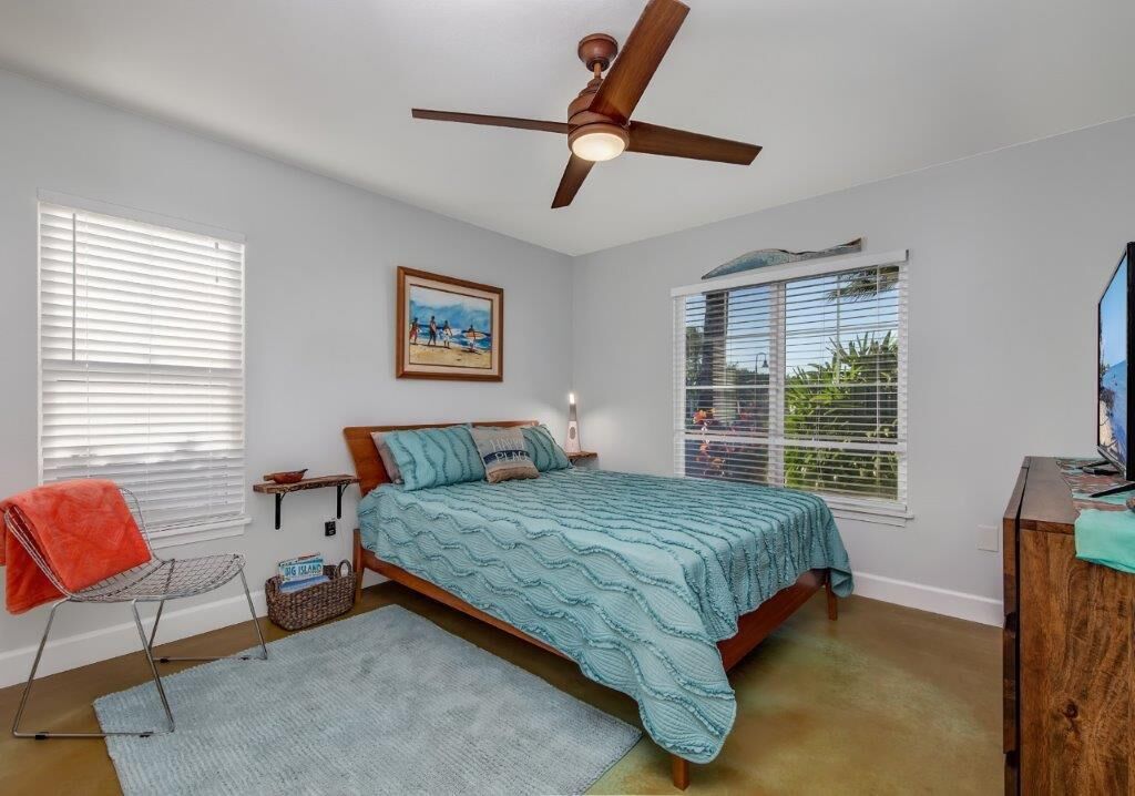 A bedroom with a bed, a ceiling fan, a window with blinds, a painting on the wall, and a small table with a lamp.