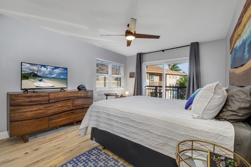 A bedroom with a bed, a wooden dresser, a ceiling fan, a TV, and a window with curtains.