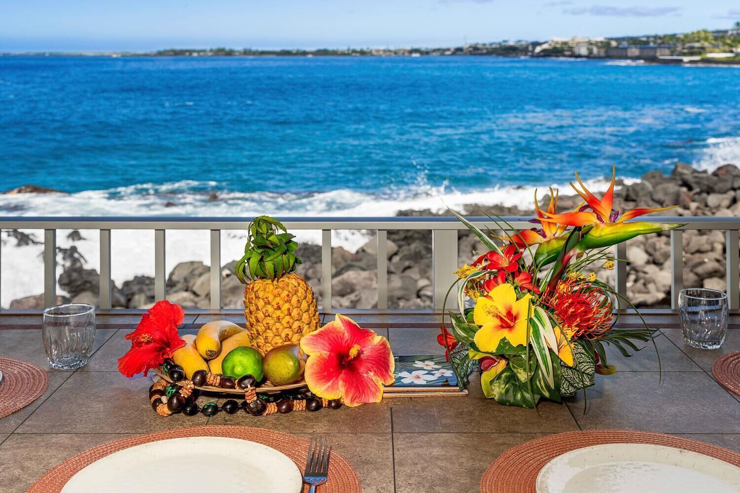 Una mesa dispuesta con frutas, flores y copas en un balcón con vistas al océano.