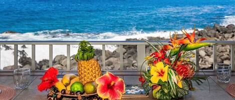 Una mesa dispuesta con frutas, flores y copas en un balcón con vistas al océano.