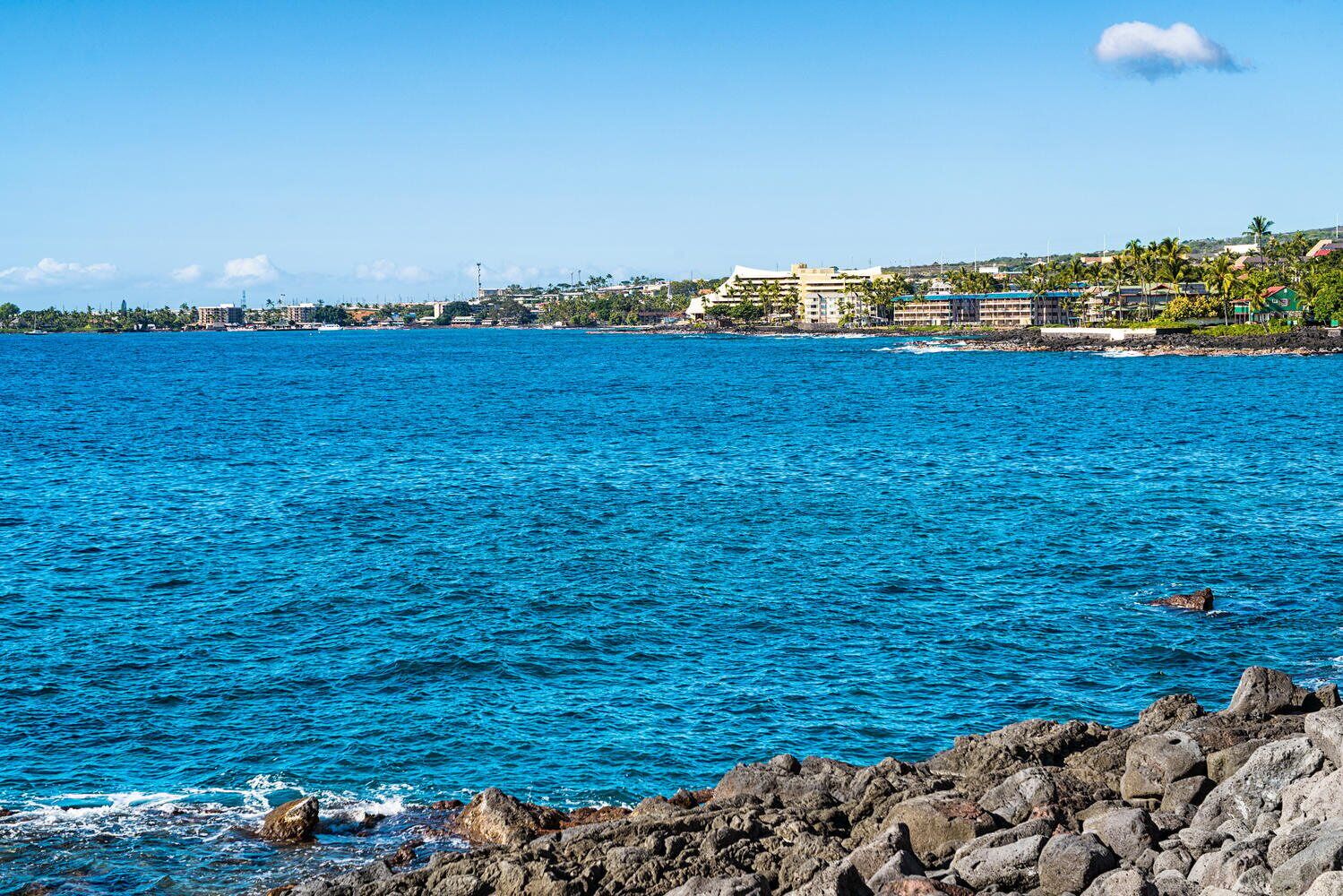 A coastal view with clear blue waters, rocky shoreline, and buildings in the background.