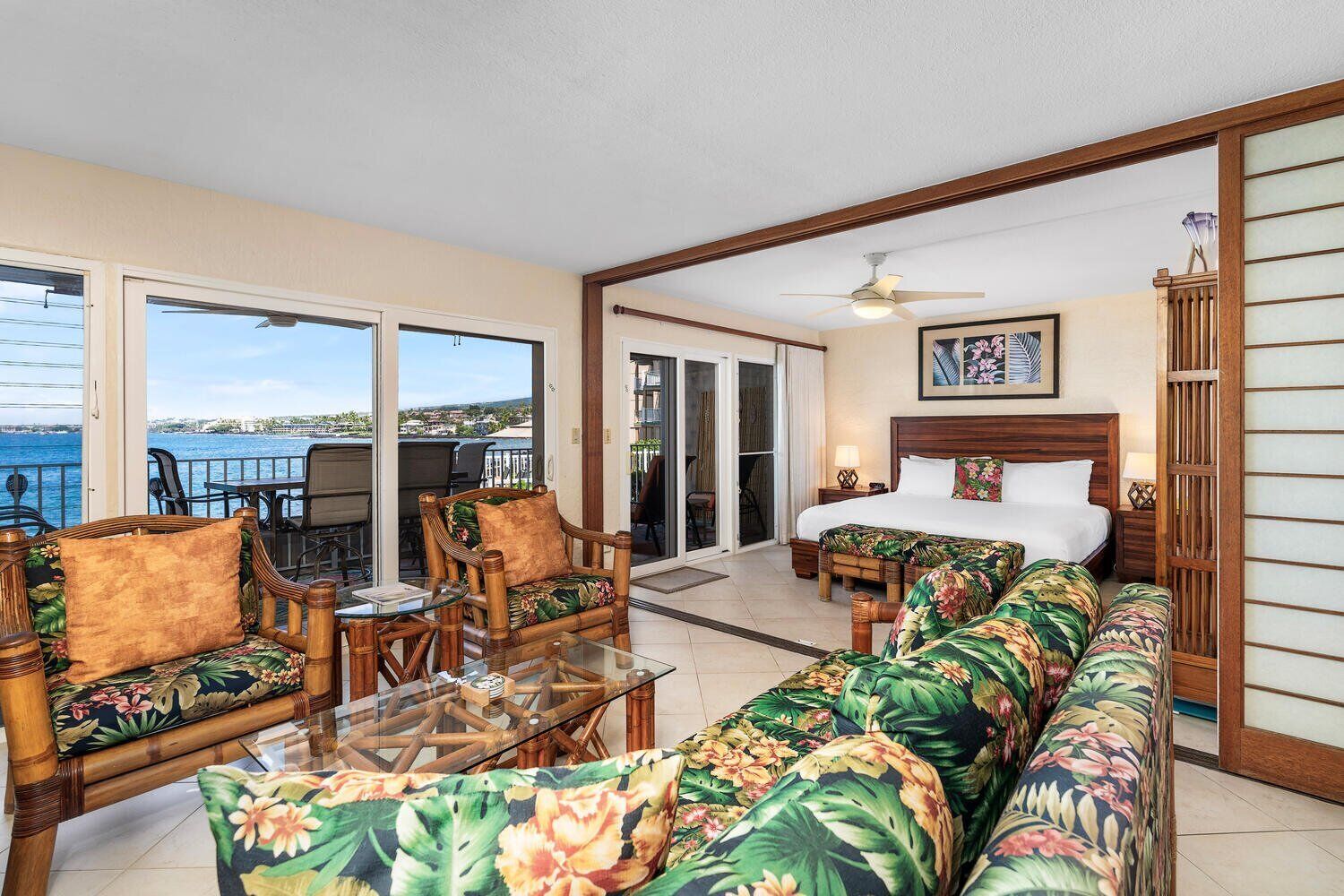 A hotel room with a bed, a sofa, a glass coffee table, and a chair. There is a view of the ocean through the windows.