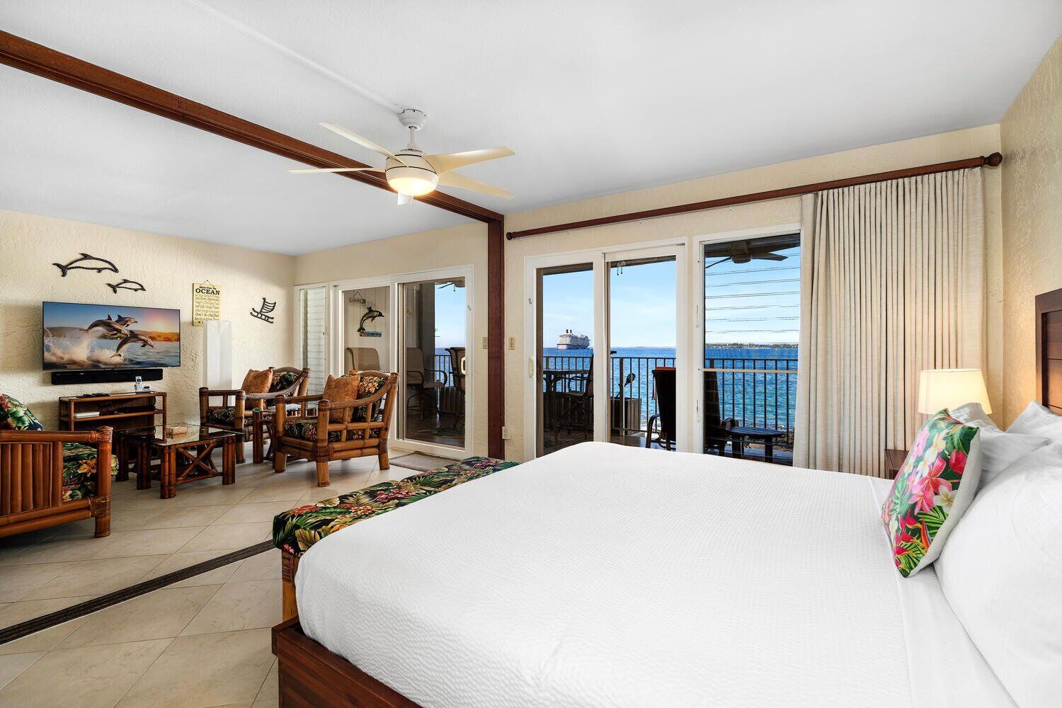 A hotel room with a large bed, a desk with chairs, a television, and a balcony with a view of the ocean.