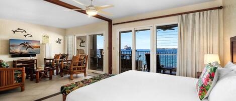 A hotel room with a large bed, a desk with chairs, a television, and a balcony with a view of the ocean.